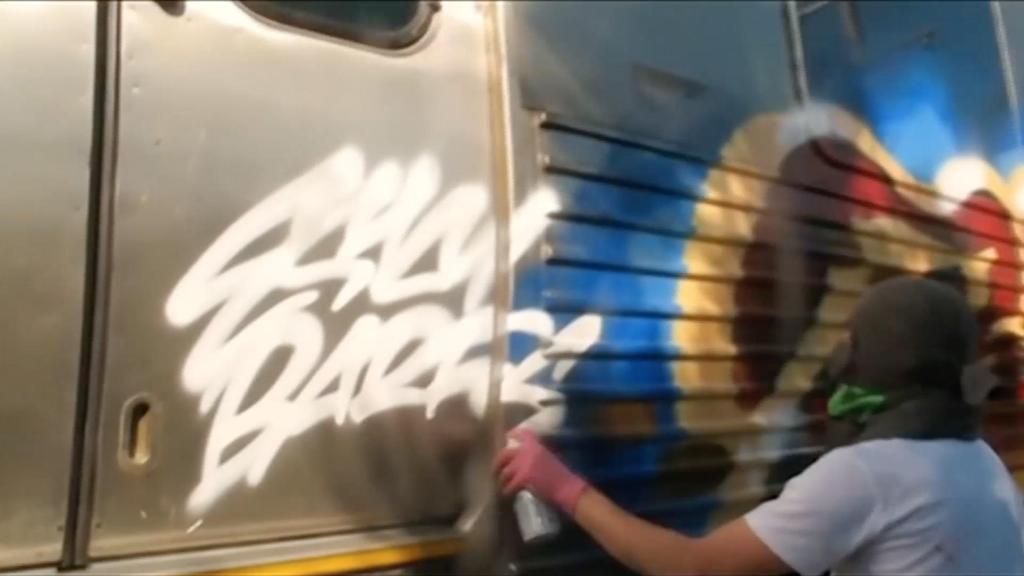 image of a hooded person spray painting words the side of a subway car at night